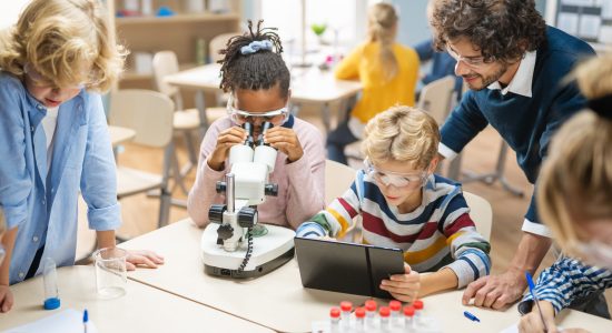 Elementary School Science Classroom: Cute Little Girl Looks Under Microscope, Boy Uses Digital Tablet Computer to Check Information on the Internet. Teacher Observes from Behind